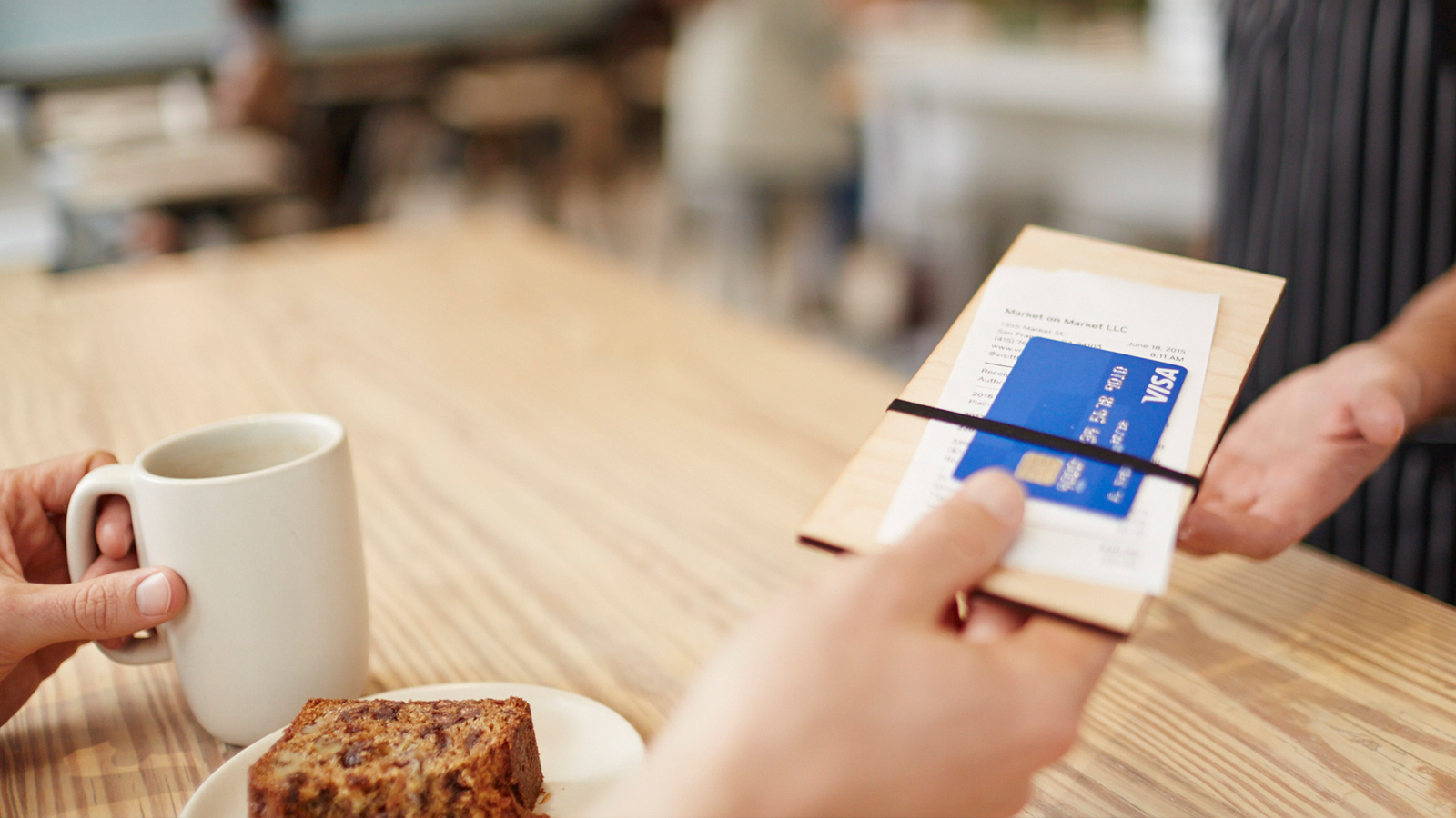 A customer sitting at a table with a mug and pastry handing the waiter the check with a Visa card.