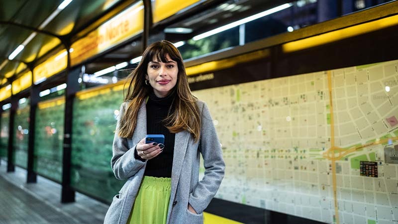 Mujer de pelo largo y castaño esperando el bus con el celular en la mano
