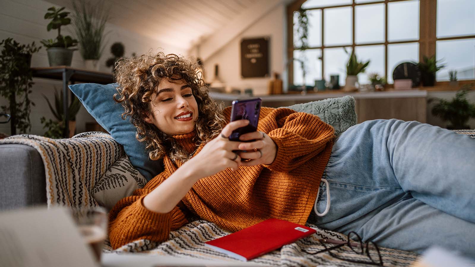 Mujer de rulos sonriente recostada en el sillón de su casa usando el celular